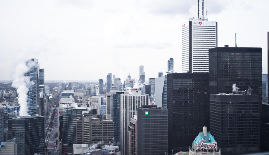 Panoramic view of downtown Toronto’s Financial District, featuring high-rise office buildings, visible steam from rooftops, and the iconic Fairmont Royal York Hotel in the foreground beneath a cloudy sky.